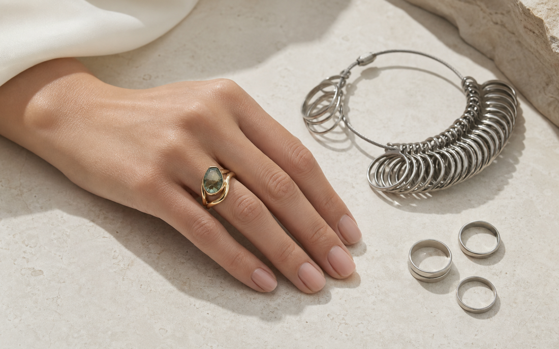 Hand wearing a sculptural gemstone ring beside metal ring-sizing gauges on a pale stone surface.